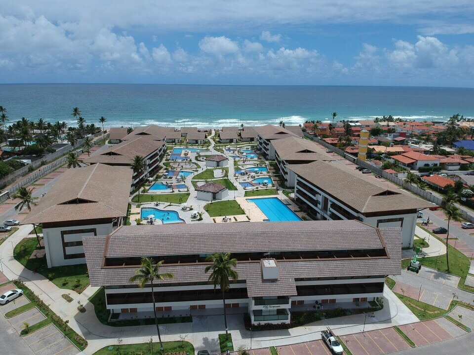 An aerial view of a coastal resort showcases multiple swimming pools surrounded by landscaped pathways. The complex features distinct buildings arranged around the pools, with palm trees lining the grounds. The ocean is visible in the background, offering a serene backdrop to the property.