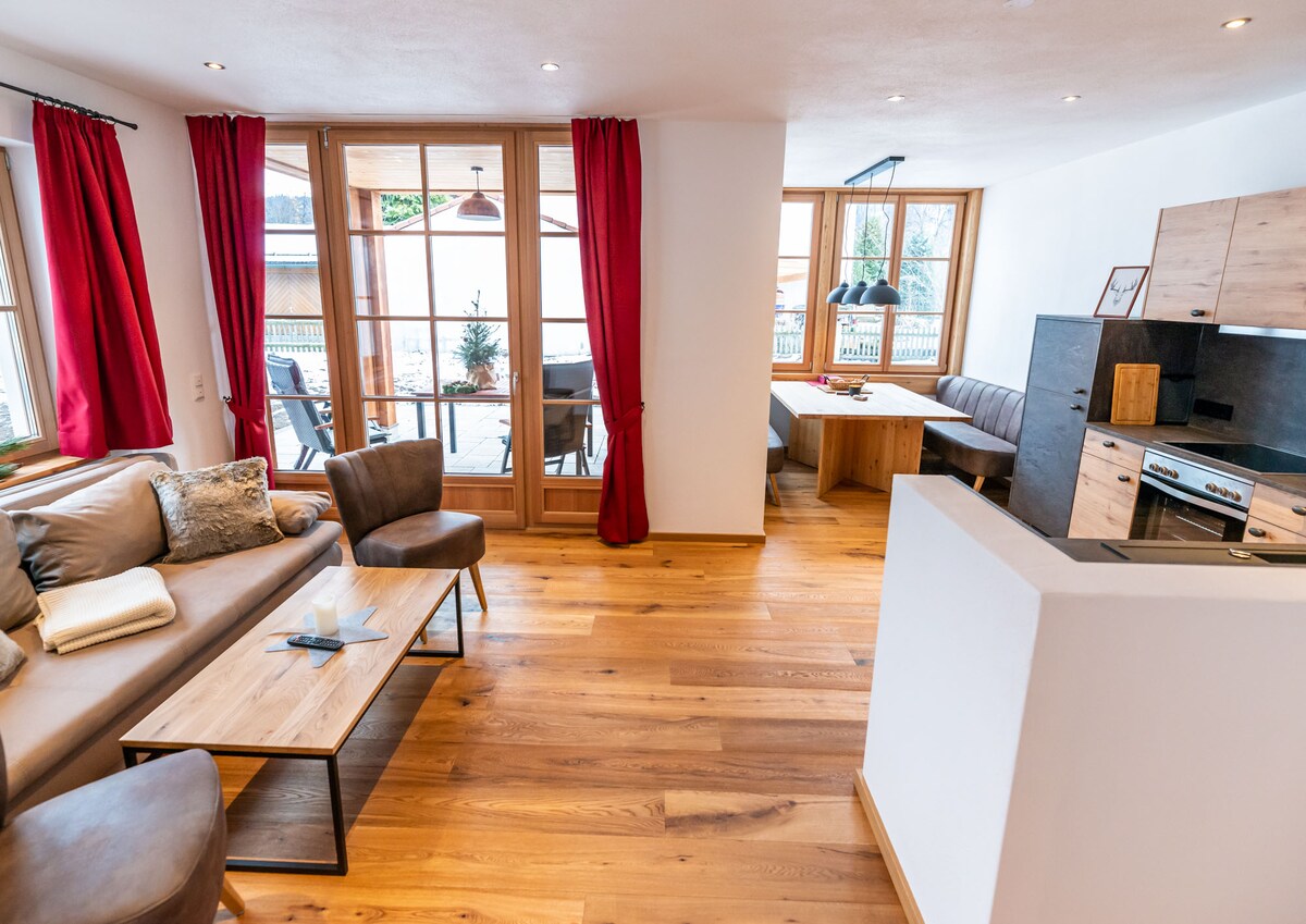 The living area features a spacious sofa accompanied by two chairs, with a wooden coffee table at its center. Large windows with red drapes provide natural light, while a dining area for up to eight people is visible. The open kitchen includes modern appliances.