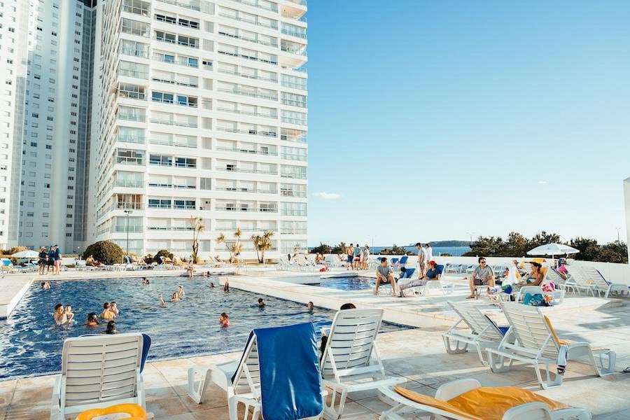 A large pool area is surrounded by a spacious patio with numerous sun loungers in white and yellow. Families and individuals are seen enjoying the water and relaxing under the sun, with a high-rise building in the background and a clear blue sky above.
