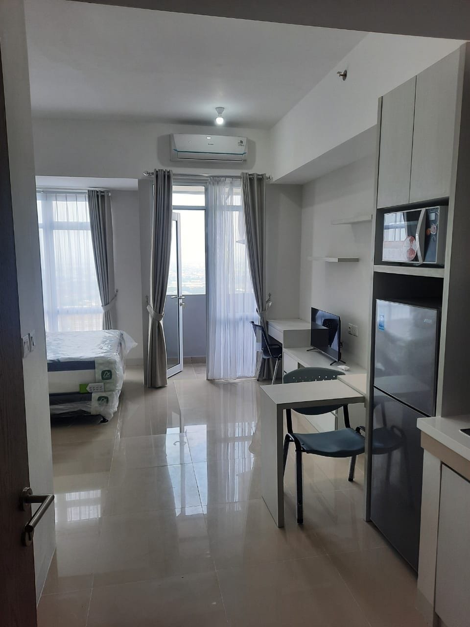 A bright and airy apartment room is visible, featuring large windows adorned with sheer curtains. A cozy sleeping area is situated to the left, while a compact dining table and chair set are positioned in the center. A kitchenette with a refrigerator and microwave is also present.