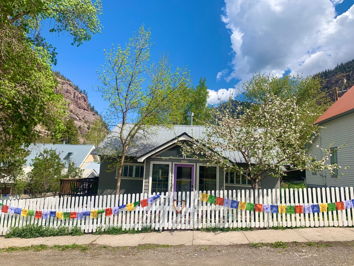 The exterior of a cozy mountain home is displayed, framed by blooming trees and colorful prayer flags. A welcoming front porch is shown, with a light-colored picket fence outlining the property. The backdrop features scenic hills under a partly cloudy sky.