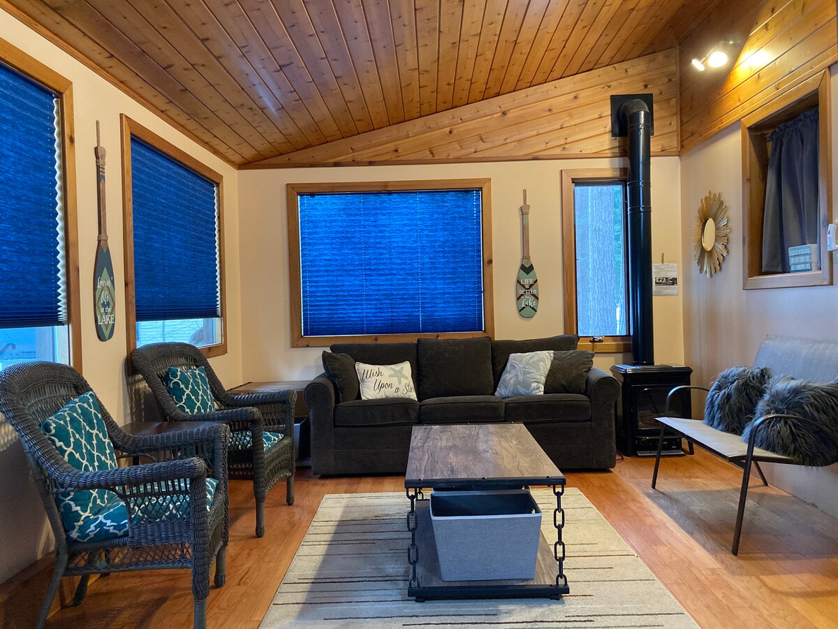 The living area features a wooden ceiling and light-colored walls, creating a spacious feel. A dark sofa is positioned centrally, accompanied by two wicker chairs. A wooden coffee table sits on a striped rug, with blue shades covering the windows, allowing for natural light.