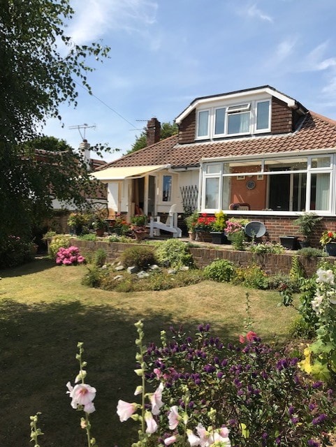 A detached house surrounded by a well-maintained garden featuring various flower beds. The front porch is visible, complemented by large windows that allow natural light into the living space. The exterior showcases a welcoming entrance with a mix of greenery and colorful blooms.