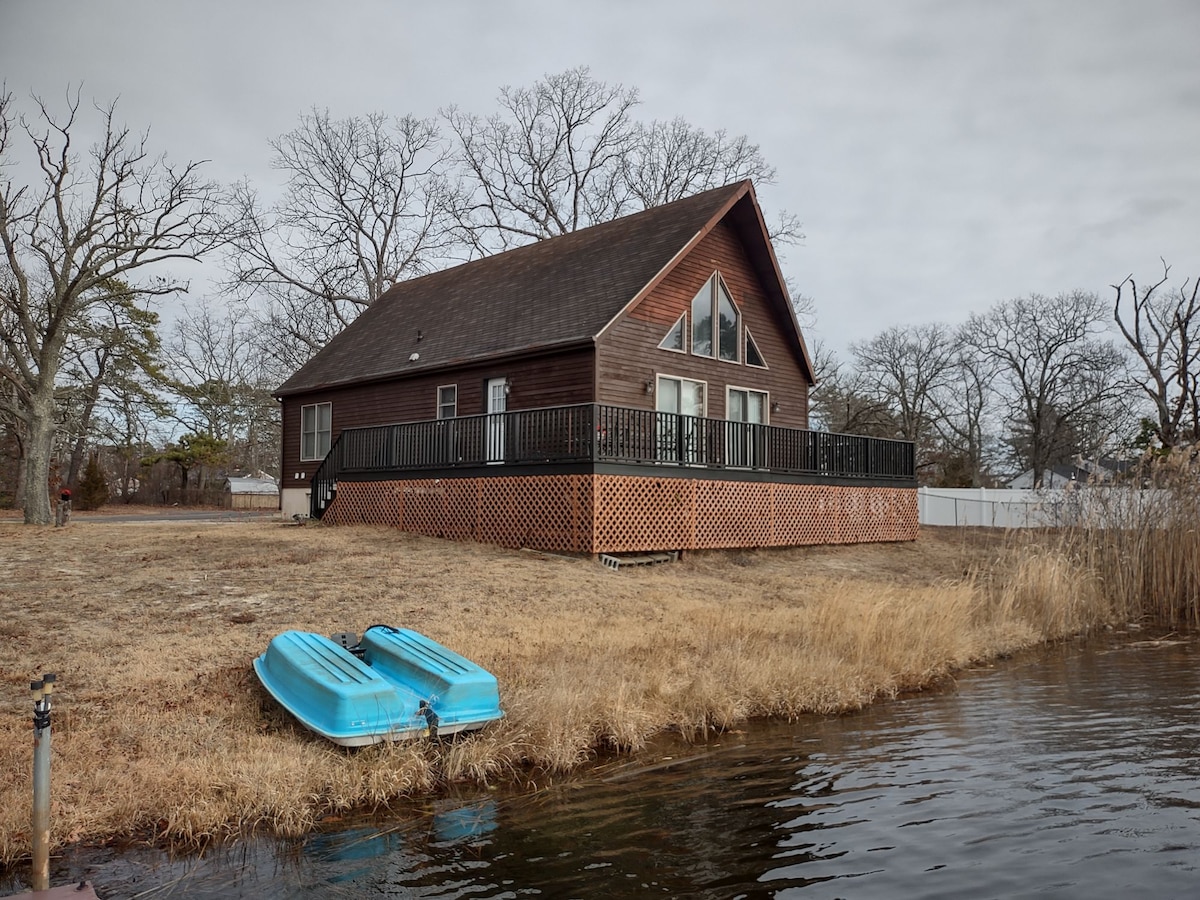 A spacious lakeside chalet is depicted with a dark wooden exterior and large windows. A wrap-around deck provides outdoor space, while two blue paddle boats rest on the grassy bank near the water, surrounded by bare trees and calm lake waters.