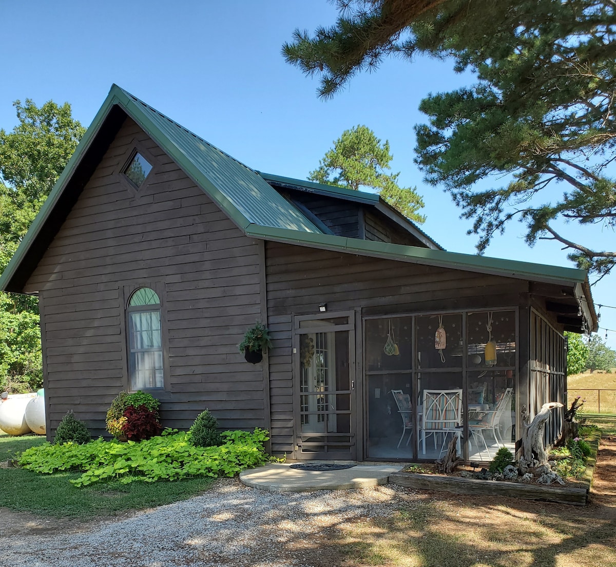 The exterior of a log-inspired guest house is displayed, featuring a gabled roof and a screened porch with seating. Lush greenery surrounds the cabin, complemented by decorative plants. A gravel path leads to the entrance, with trees providing shade in the background.