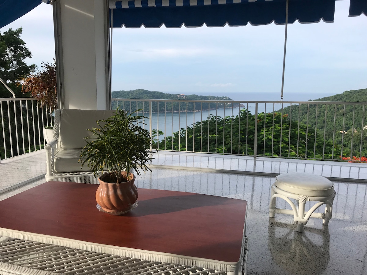 A terrace seating area features a wooden table with a potted plant at its center. White lounge chairs are positioned nearby, offering views of the lush green hills and the calm bay in the distance, under a striped awning providing shade.
