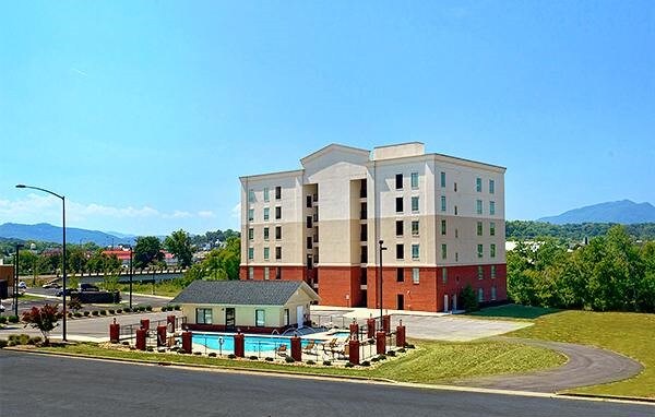A modern multi-story condo building is shown against a clear blue sky, with surrounding green areas and distant mountains. A swimming pool area with lounge chairs is visible in the front, and parking space is prominently featured.