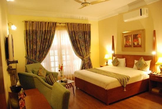 A well-lit room features a spacious bed with a coordinating throw. Soft drapes frame the large windows, allowing natural light to fill the space. A small seating area is included, with a green couch and side tables. Decorative artwork adds subtle charm to the walls.