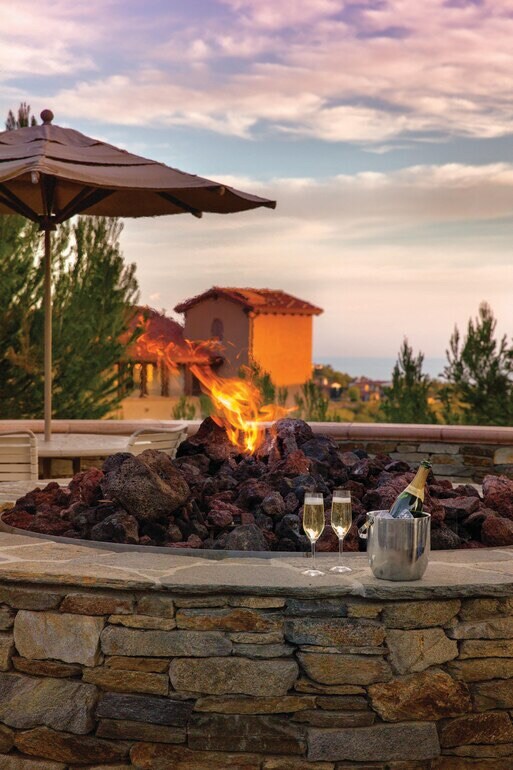 A stone fire pit is surrounded by smooth, dark rocks, with flames rising gracefully. A metal bucket containing champagne sits beside two sparkling glasses. In the background, Mediterranean-style buildings are framed by lush greenery against a colorful sky.