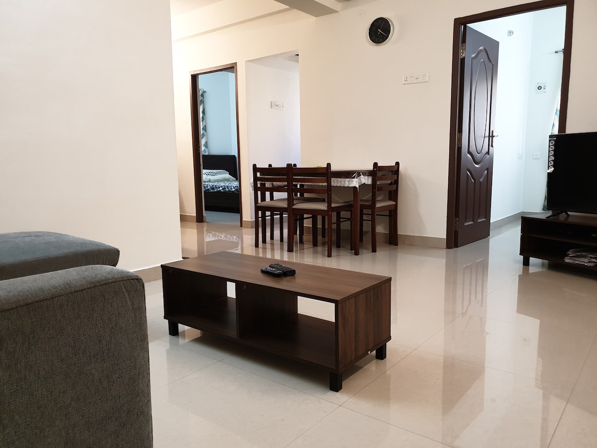 A living area is shown featuring a rectangular coffee table in front of a sofa. A dining table with four wooden chairs sits in the background, while two doorways lead to other rooms. The walls are painted in neutral tones, highlighting the spacious layout.