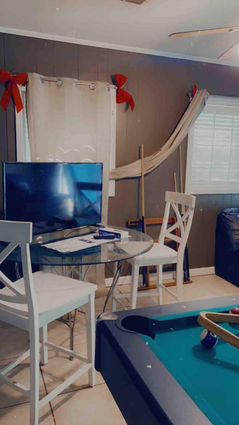 A small dining area is visible, featuring a round glass table paired with two white chairs. A television stands on a media console nearby, with a game room ambiance created by a pool table in the foreground. Decor includes two bows hanging on the wall, adding a festive touch.