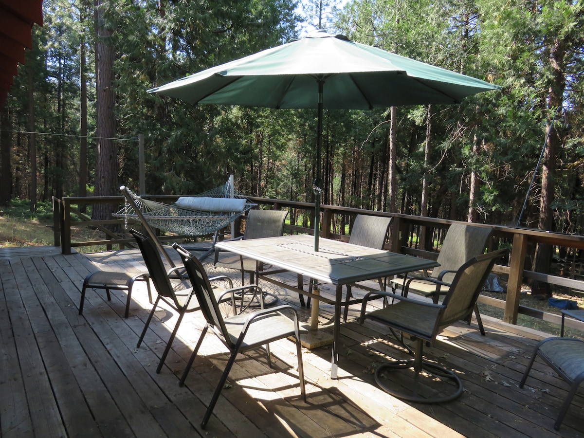 A spacious wooden deck is surrounded by tall trees, offering a shaded area with a large green umbrella. A sturdy table with several chairs is present, alongside a hammock positioned nearby, creating a comfortable outdoor space for relaxation.