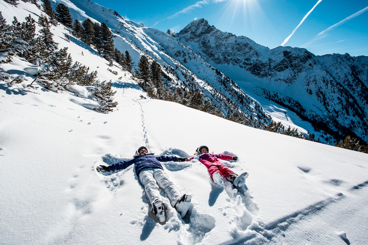 Two individuals lie in the snow, creating angels against a backdrop of a snow-covered mountain landscape. Sunlight illuminates the scene, highlighting the surrounding pine trees and the pristine snow. Fresh tracks lead away from their positions, suggesting recent activity in the area.