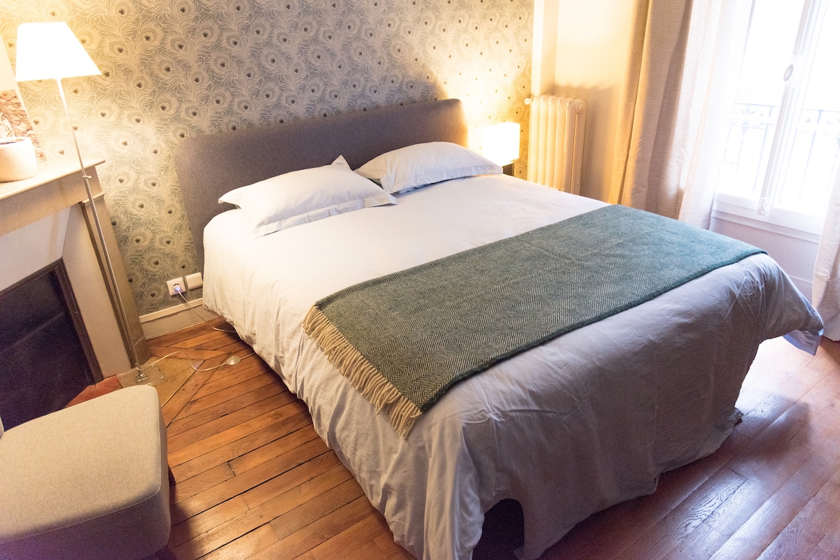 A spacious bedroom is presented with a comfortable bed adorned with a light blanket. Soft lighting is provided by a bedside lamp, and a unique patterned wallpaper adds subtle character. Wooden flooring contributes warmth to the room, while natural light filters in through a nearby window.