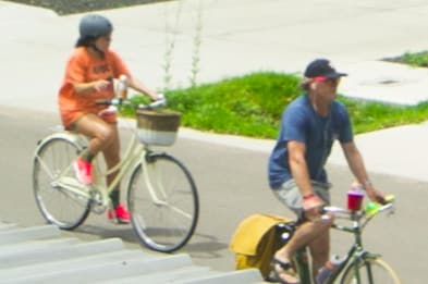 Two individuals are riding bicycles along a paved path. One cyclist wears an orange shirt and rides a light-colored bike with a front basket. The other cyclist is dressed in a blue shirt and is on a dark bike with a bag attached, both enjoying a sunny day.