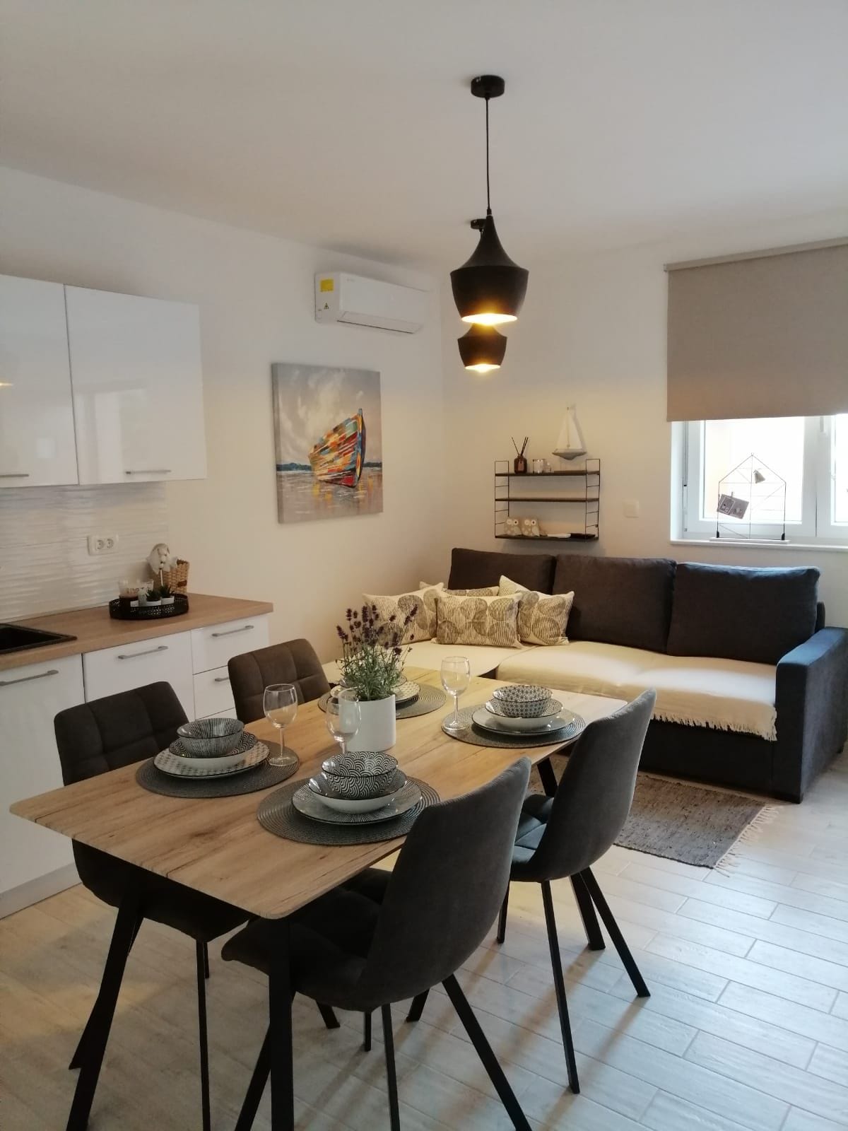 A modern dining area is paired with a cozy living space. A table is set for four, featuring neatly arranged plates and glasses. A dark sofa with decorative pillows complements the light cabinetry. A wall art piece adds a splash of color, while natural light filters through a window.