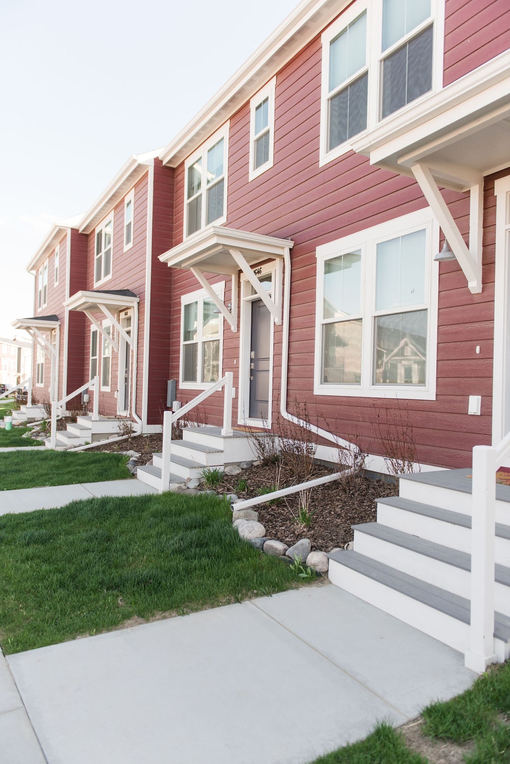 A row of modern townhomes features a uniform facade of rich red siding. Each home has a welcoming front porch with white railings and steps leading up to the entrance. Neatly landscaped grass and decorative stone borders enhance the appealing exterior.