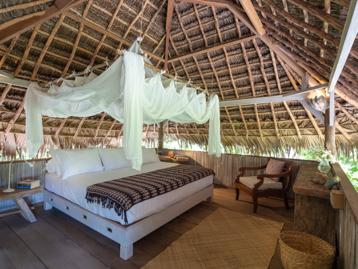 An open-air bungalow features a spacious bed with white linens and a decorative woven blanket. The thatched roof, made from natural materials, creates a warm ambiance. A wooden chair and a rustic table complement the relaxing atmosphere, while soft light filters through the space.