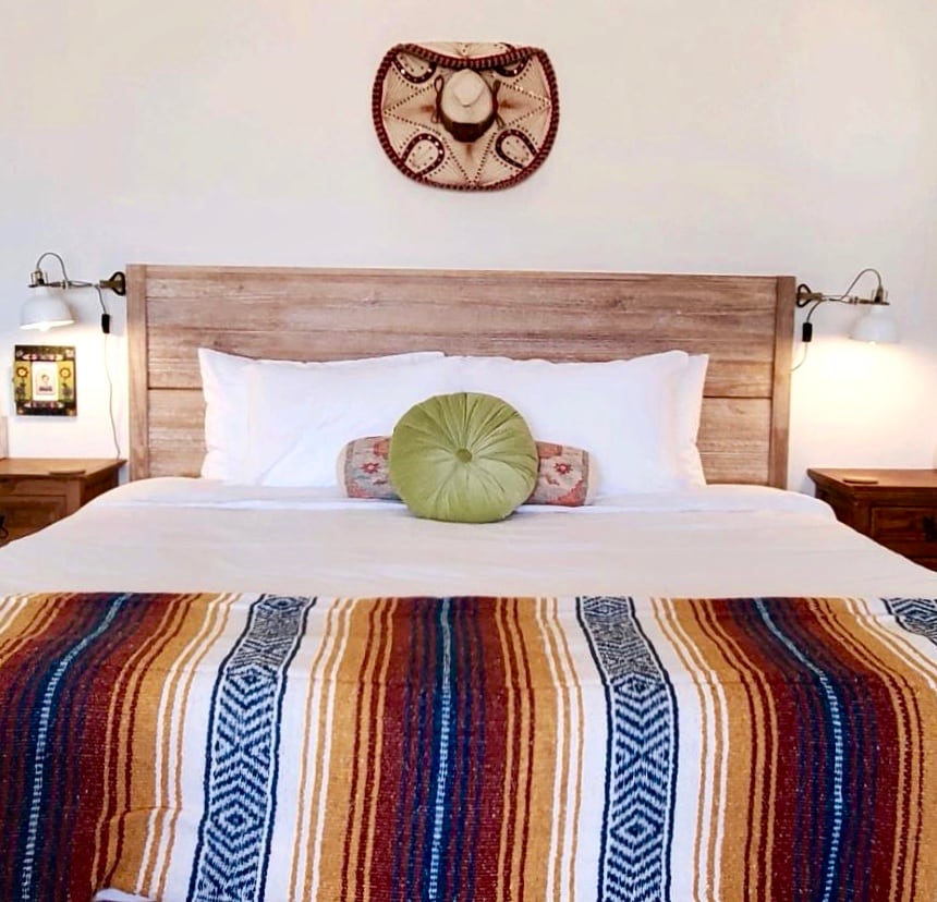 A well-appointed sleeping area features a spacious bed adorned with a colorful woven blanket and a decorative pillow. Table lamps flank the bed, providing soft lighting. A decorative wall piece hangs above the headboard, adding a touch of local charm.