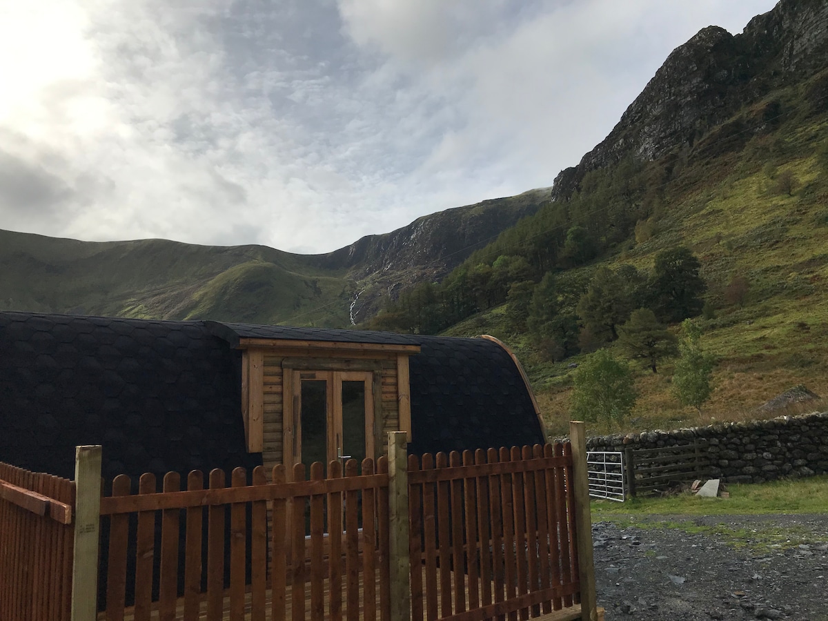 A wooden hut is positioned within a mountainous landscape, featuring a fenced area. The structure displays a rounded roof and a large window, providing views of the surrounding hills and distant waterfall. Lush greenery and rocky terrain complement the serene setting.