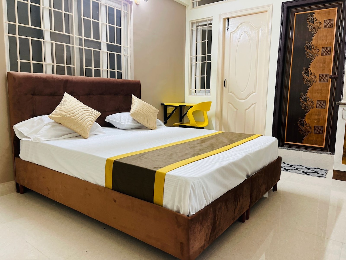 A spacious sleeping area is furnished with a soft brown bed featuring a yellow and brown bedspread. Decorative pillows are arranged on the bed. The room is illuminated by natural light filtering through multiple large windows, and a modern entrance door adds character.