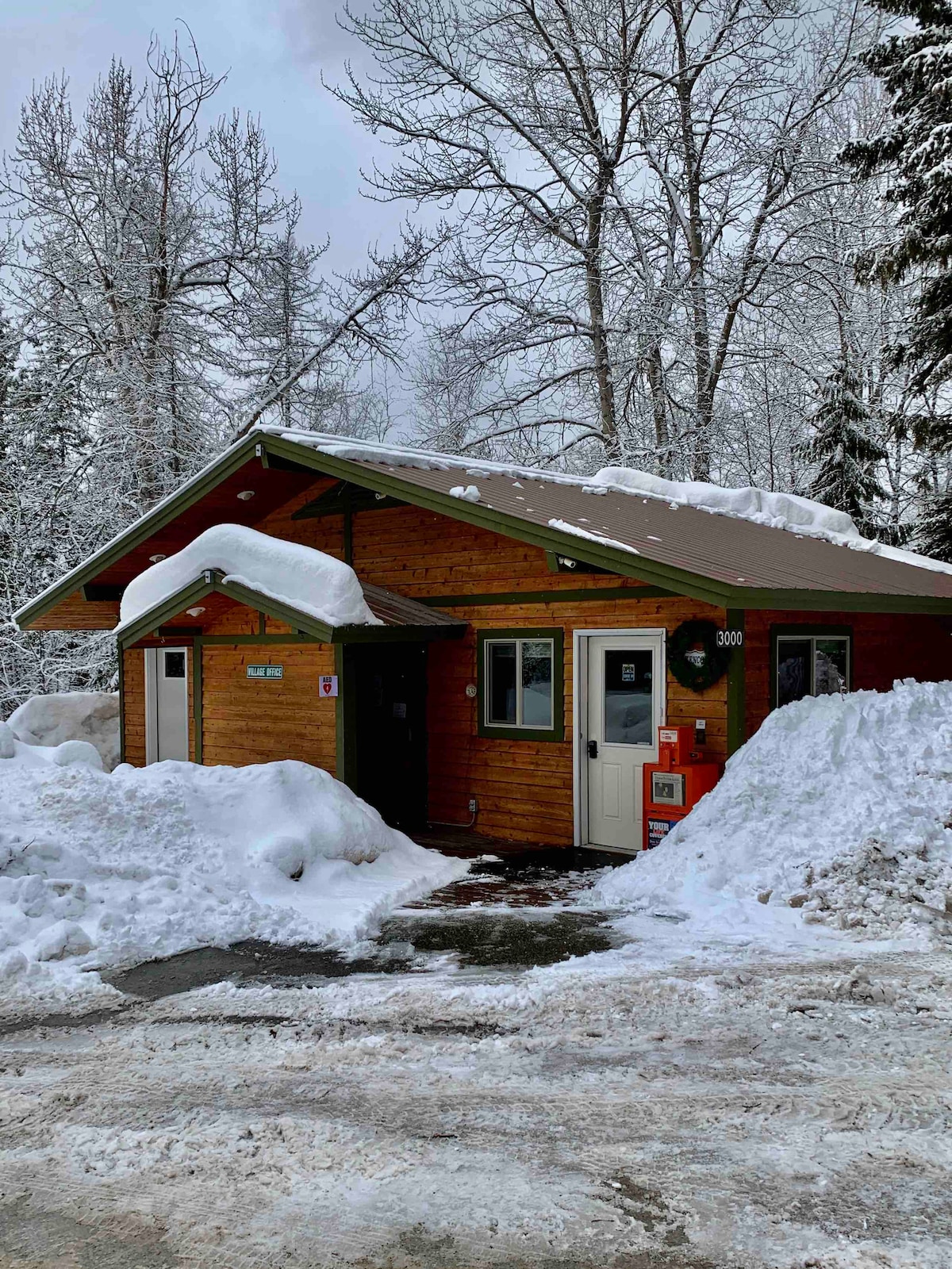 The exterior of a wooden triplex unit is shown, set against a snowy landscape. The angled roof is covered with snow, and the entrance features a welcoming porch with a festive wreath. Snow piles are seen surrounding the building, indicating a winter setting.