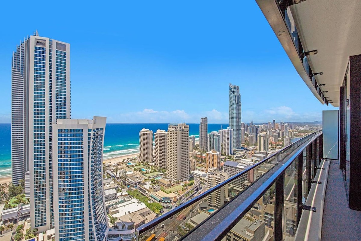 A sweeping view of the Pacific Ocean and Surfers Paradise showcases a vibrant coastline and high-rise buildings. The expansive balcony offers a clear vantage point, with glass railings ensuring unobstructed sightlines. The bright blue sky contrasts with the urban landscape, enhancing the scene's appeal.