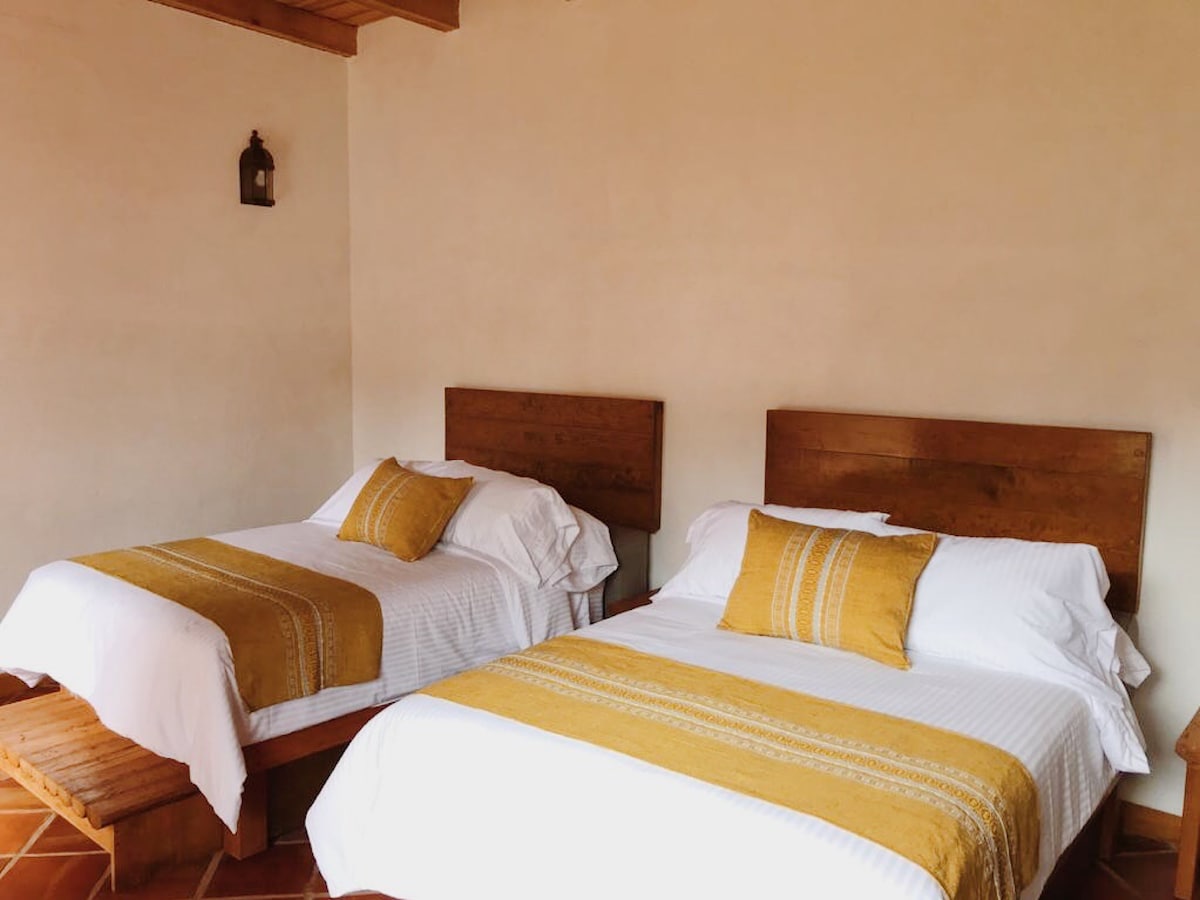 A cozy sleeping area features two spacious beds with soft white linens and yellow patterned throws. Simple wooden headboards complement the warm tones of the terracotta tiled floor, while soft natural light fills the room, creating a calming atmosphere.