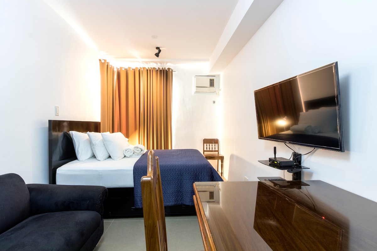 A well-lit room features a spacious bed with neatly arranged white linens and a navy blue blanket. A television is mounted on the wall opposite a wooden table and chair. Dark curtains frame a window, and a cozy black sofa adds to the inviting ambiance.