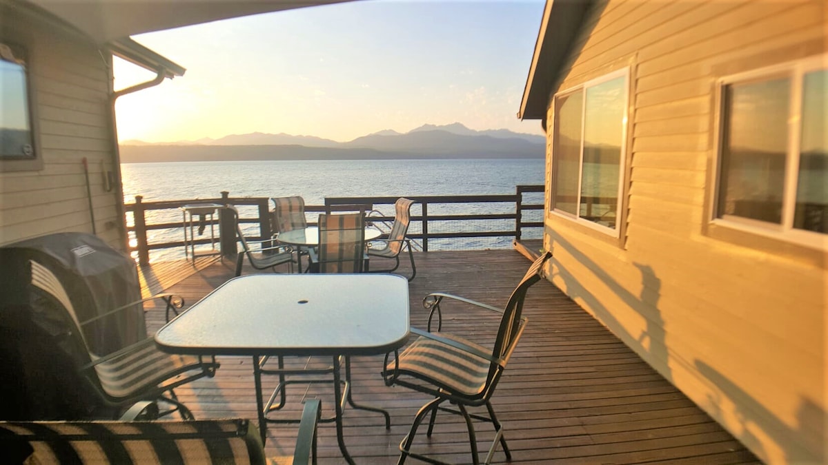 A spacious deck offers stunning views of the water and mountains at sunset. The area features a glass-top table surrounded by striped outdoor chairs, along with a Weber gas grill positioned nearby. The warm light enhances the inviting setting.