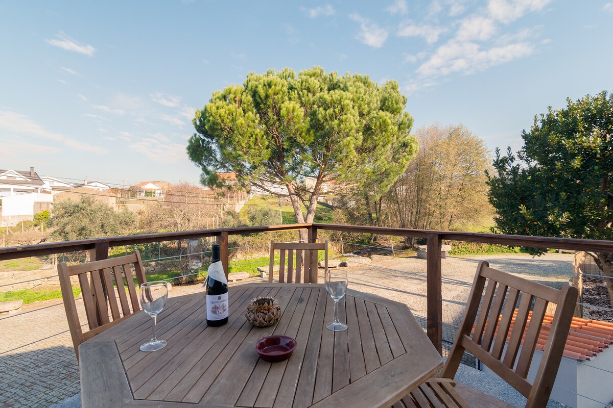 An outdoor seating area features a round wooden table with four matching chairs. A bottle of wine and glasses are placed alongside a small dish of snacks. A large tree provides shade, and a garden landscape is visible in the background.