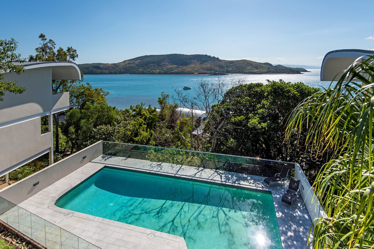A sparkling swimming pool is featured in the foreground, offering views of tranquil waters and lush greenery. Beyond the pool, the gentle curves of the landscape lead to a mountainous backdrop, enhancing the sense of serenity and connection to nature.