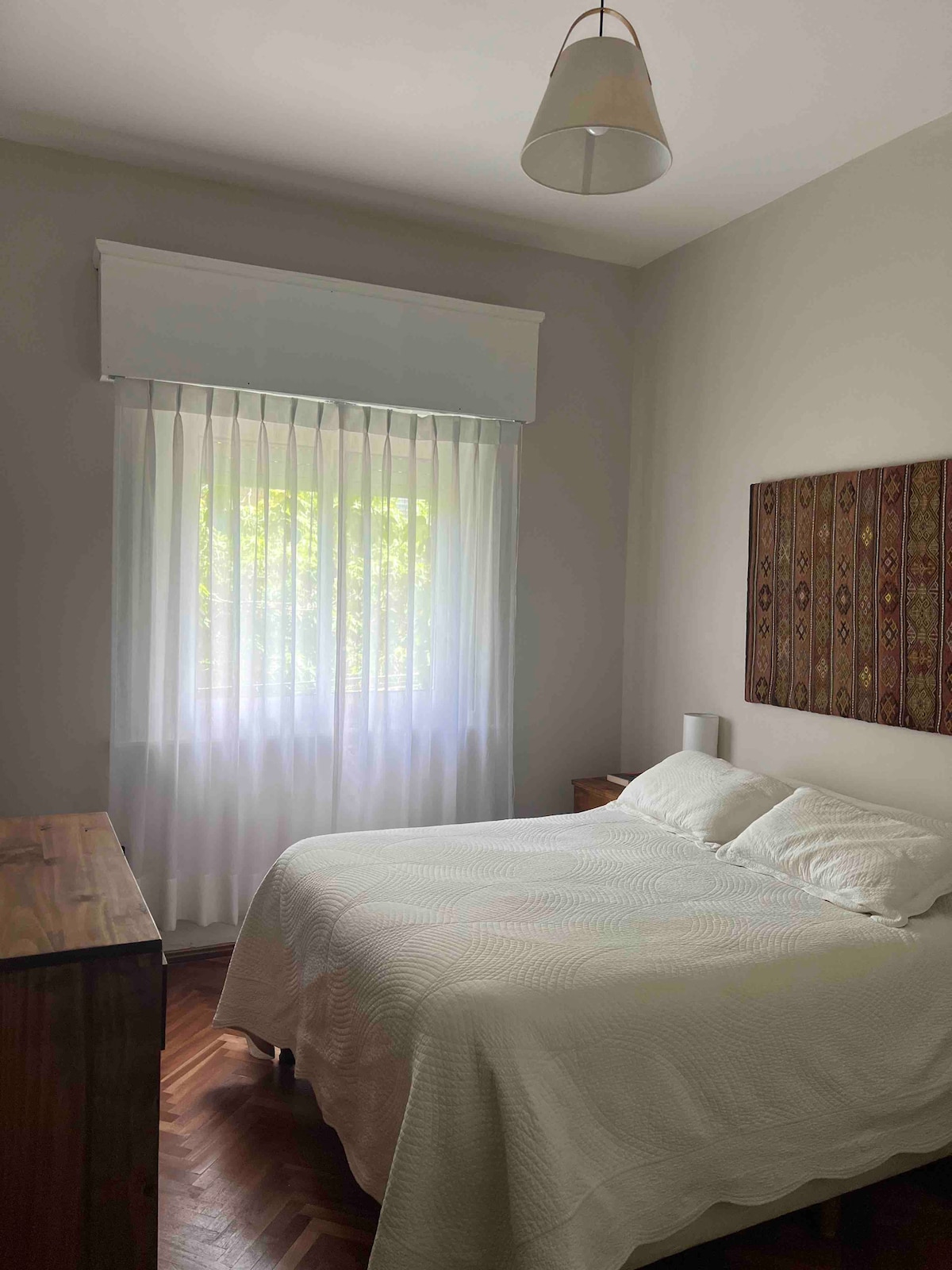 The serene bedroom features a spacious bed with a light-colored coverlet, complemented by soft natural light filtering through sheer white curtains. A wooden nightstand and dresser offer functional storage, while a patterned wall art piece adds subtle texture to the room's neutral tones.