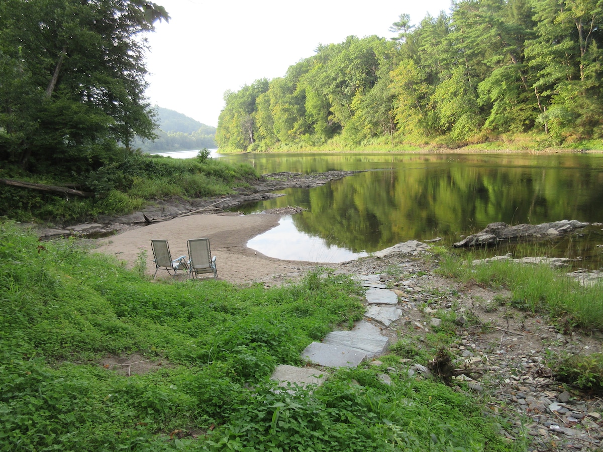 A sandy beach area by the river is framed by lush green vegetation. Two lounge chairs are positioned on the sand, facing the water. The calm river reflects the surrounding trees, creating a serene natural setting.