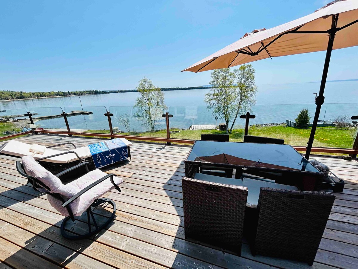 A spacious deck overlooks Lake Superior, featuring a shaded patio umbrella and outdoor seating. Comfortable lounge chairs and a table provide a relaxing space to enjoy the serene water views. The natural surroundings are complemented by lush greenery and a dock in the distance.