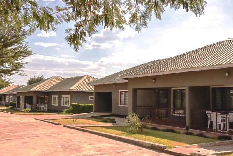 Multiple serviced villas are arranged along a paved path, each with a distinct entrance and a shaded porch. The properties feature well-maintained gardens and a clear blue sky with soft clouds overhead, highlighting the tranquil setting.