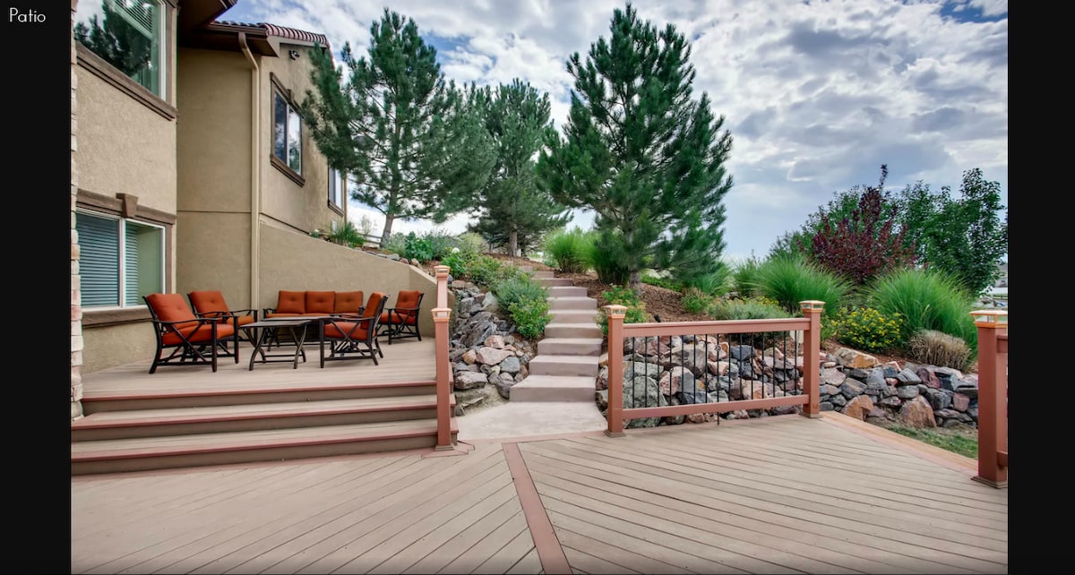 A spacious outdoor patio features a seating area with multiple chairs around a large table. Stone steps lead up to a lush, greenery-filled garden, enhancing the natural surroundings. The deck's wooden flooring supports a relaxed outdoor setting, perfect for enjoying the landscape.
