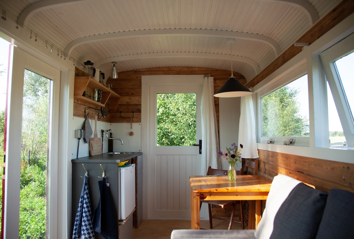 The interior of the tiny house features wooden paneling and a cozy seating area. A table is positioned beside a kitchen counter with essential appliances. Large windows provide views of the surrounding greenery, allowing natural light to enhance the space.