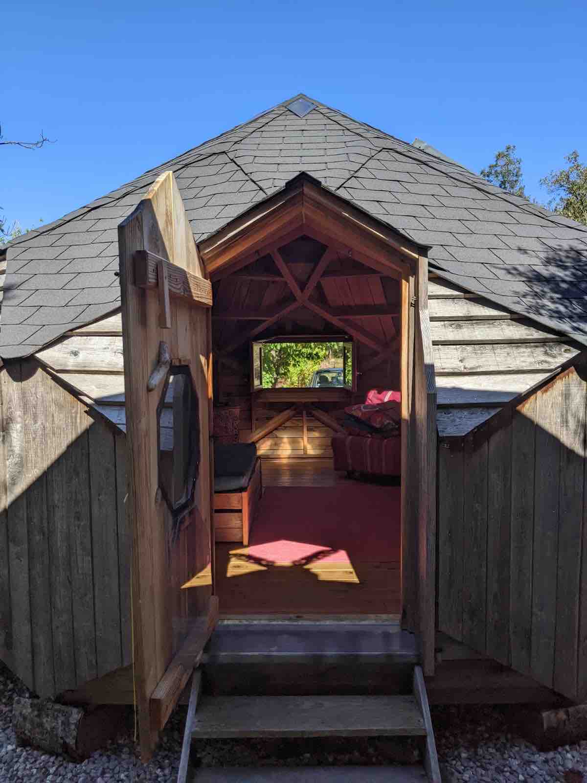 An entrance is shown leading into a wooden structure with a unique angular design. Light pours in through the open door, highlighting a warm interior with a cozy seating area visible. Surrounding greenery is seen through the windows, enhancing the sense of connection with nature.