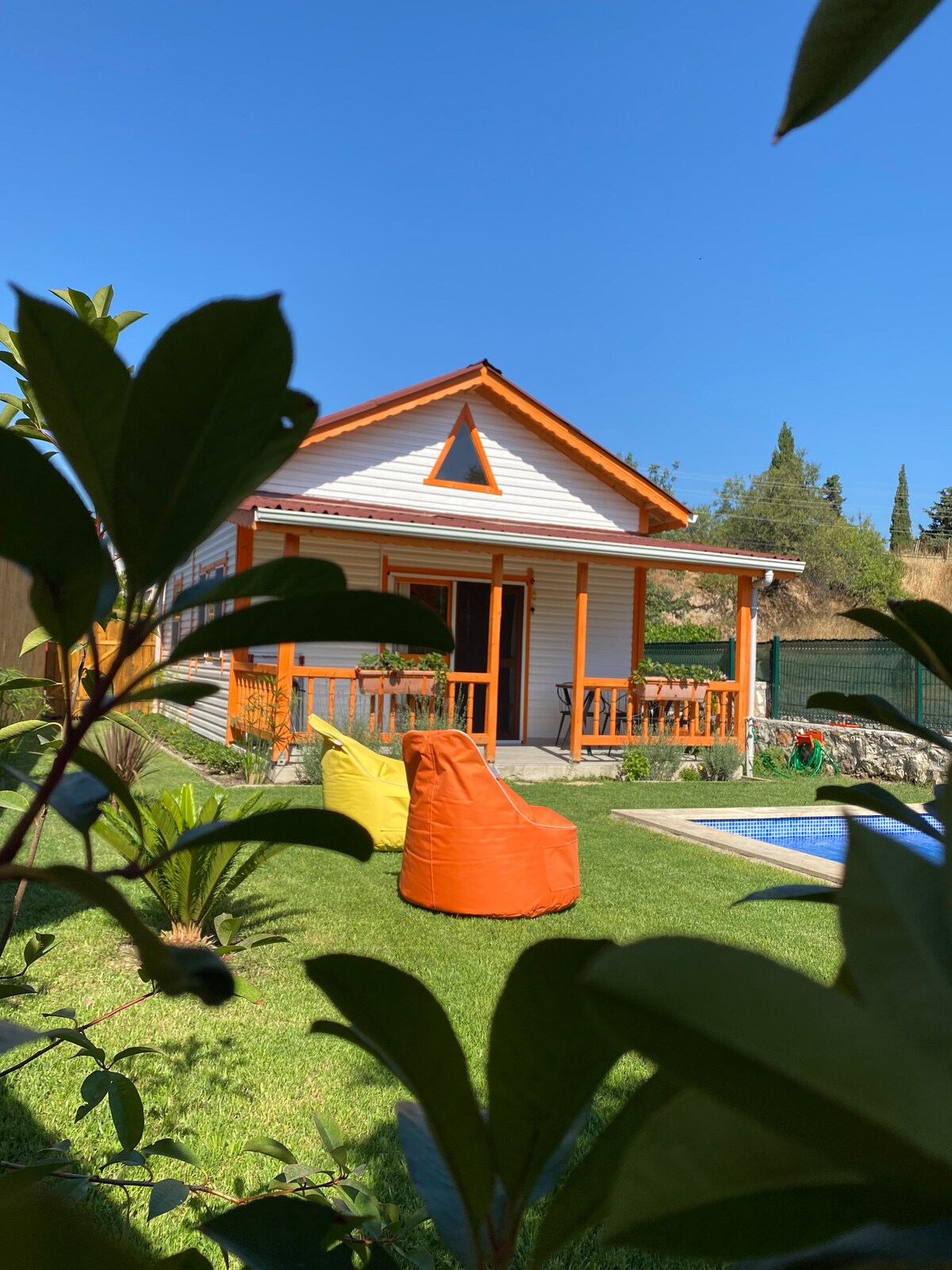 A charming bungalow is situated among lush greenery, featuring a triangular roof and a welcoming porch. Two colorful bean bag chairs are positioned on the manicured lawn, which surrounds a private swimming pool, offering a serene setting for relaxation.