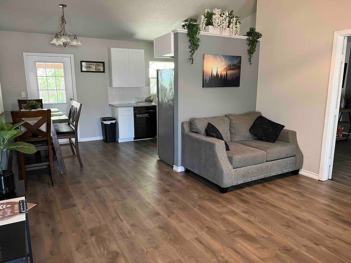 The open living space features light wood flooring and is furnished with a comfortable gray sofa. A dining area with four chairs is visible, along with a modern kitchen setup, including stainless steel appliances and a decorative wall-mounted photo. Natural light enters through the window.