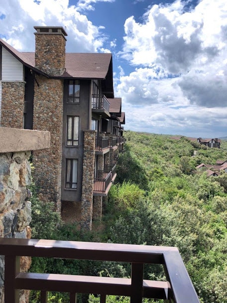 The image captures an elegant multi-story villa nestled amidst lush greenery. The stone and wood exterior is complemented by large windows on each floor. A balcony railing frames the view, while clouds drift above in the bright sky, showcasing the natural beauty of the surrounding landscape.