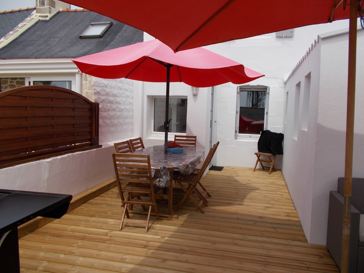 A spacious terrace is pictured, featuring a wooden deck and a large table surrounded by folding chairs. Red umbrellas provide shade, while a barbecue is positioned nearby. The white walls create a bright, airy environment, contributing to an outdoor dining space.