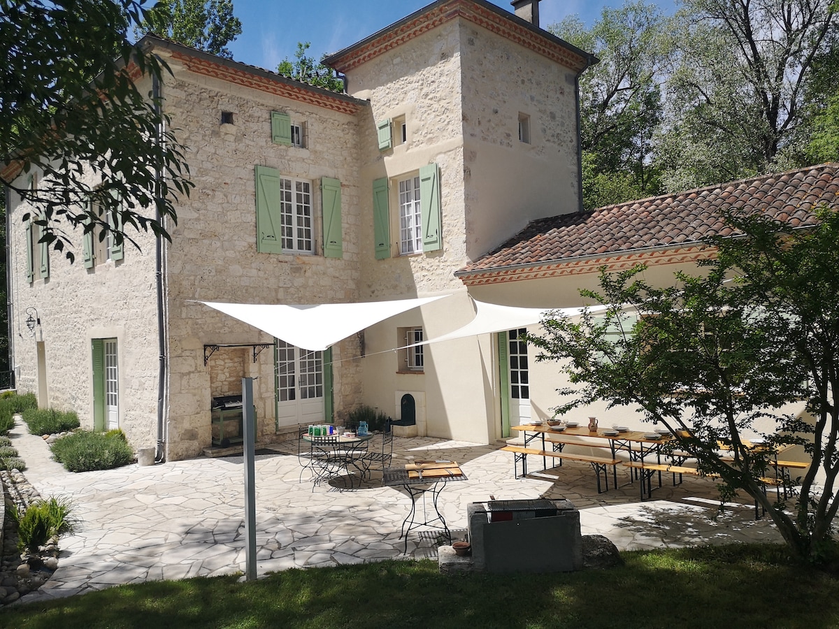 A charming exterior view of the restored mill features a stone facade with green shutters. Sunshades provide shelter over an outdoor seating area, while a large dining table accommodates multiple guests. Lush greenery surrounds the property, enhancing its natural setting.