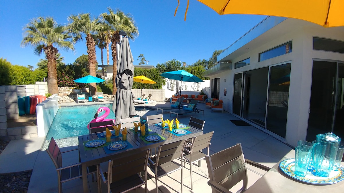 An outdoor dining area is set with colorful plates and napkins, offering a view of the inviting pool surrounded by lounge chairs and vibrant umbrellas. Tall palm trees create a tropical atmosphere, while a shaded area features a large umbrella for comfort.