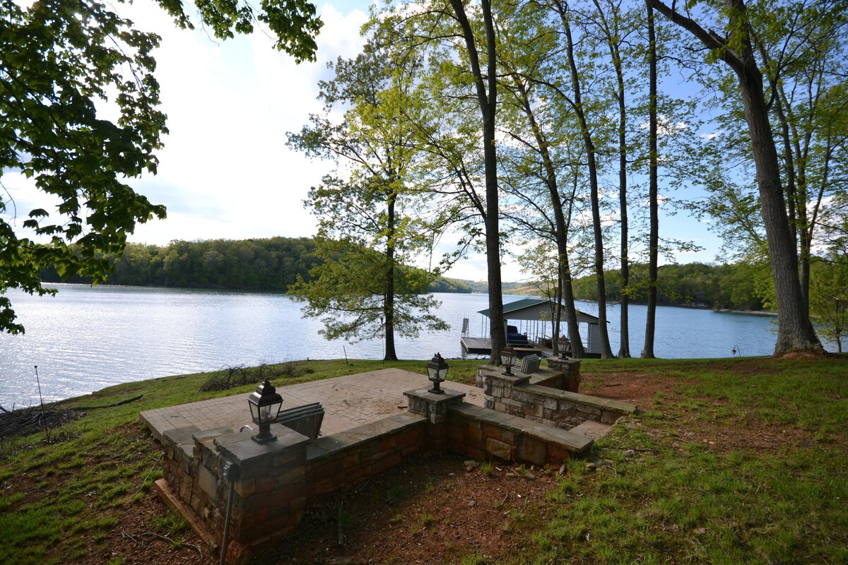 A serene outdoor space is set near the water's edge, featuring a stone patio surrounded by trees. Four lanterns light the area, providing soft illumination. The expansive lake view is visible in the distance, framed by lush greenery and gentle hills.