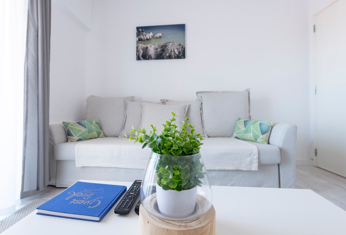 A light-filled living area showcases a soft, neutral-colored sofa with decorative cushions. A small round table is accented with a potted plant and a book, while a remote control sits nearby. A framed image of a coastal scene adds a subtle artistic touch to the wall.