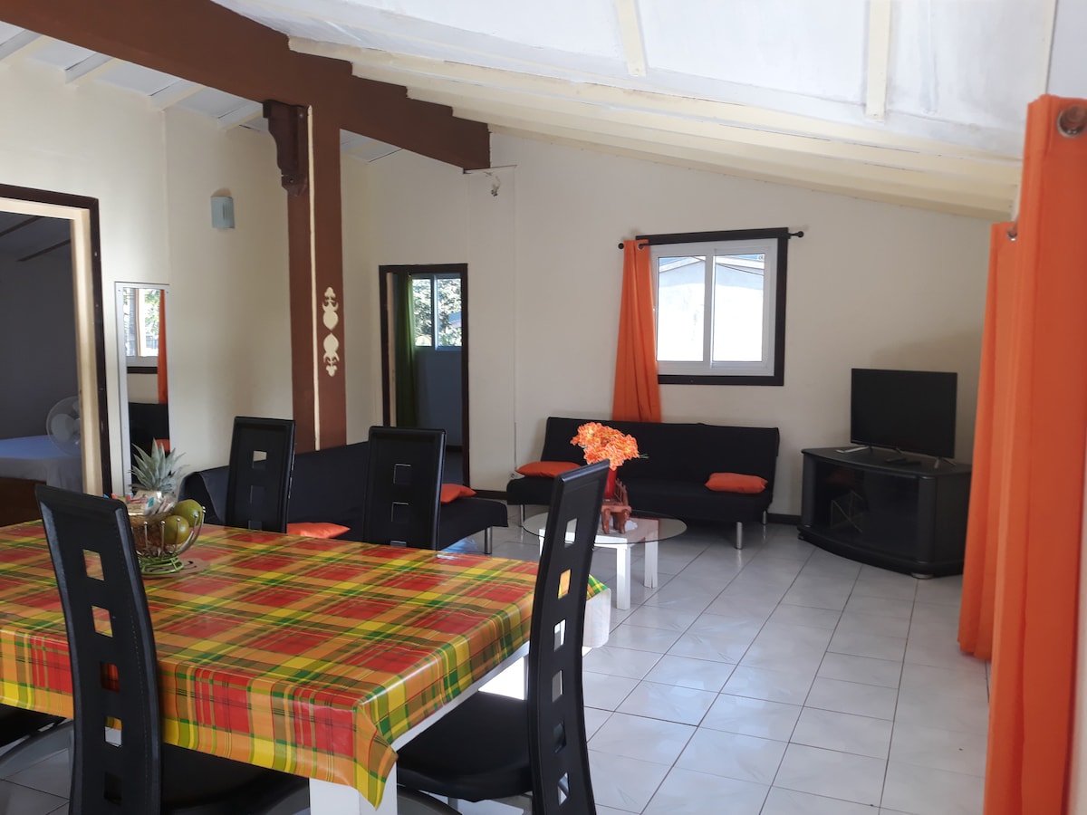 A bright living area features a dining table with a colorful checkered tablecloth. Comfortable seating is arranged around a television, with lively orange curtains framing the windows. Light reflects off the tiled floor, enhancing the spacious feel of the room.