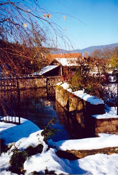 A serene winter landscape is captured with a gently flowing stream bordered by snow-covered ground. Stone walls and greenery partially frame the scene, while a house with a sloped roof is visible in the background. Bright blue skies complete the tranquil setting.