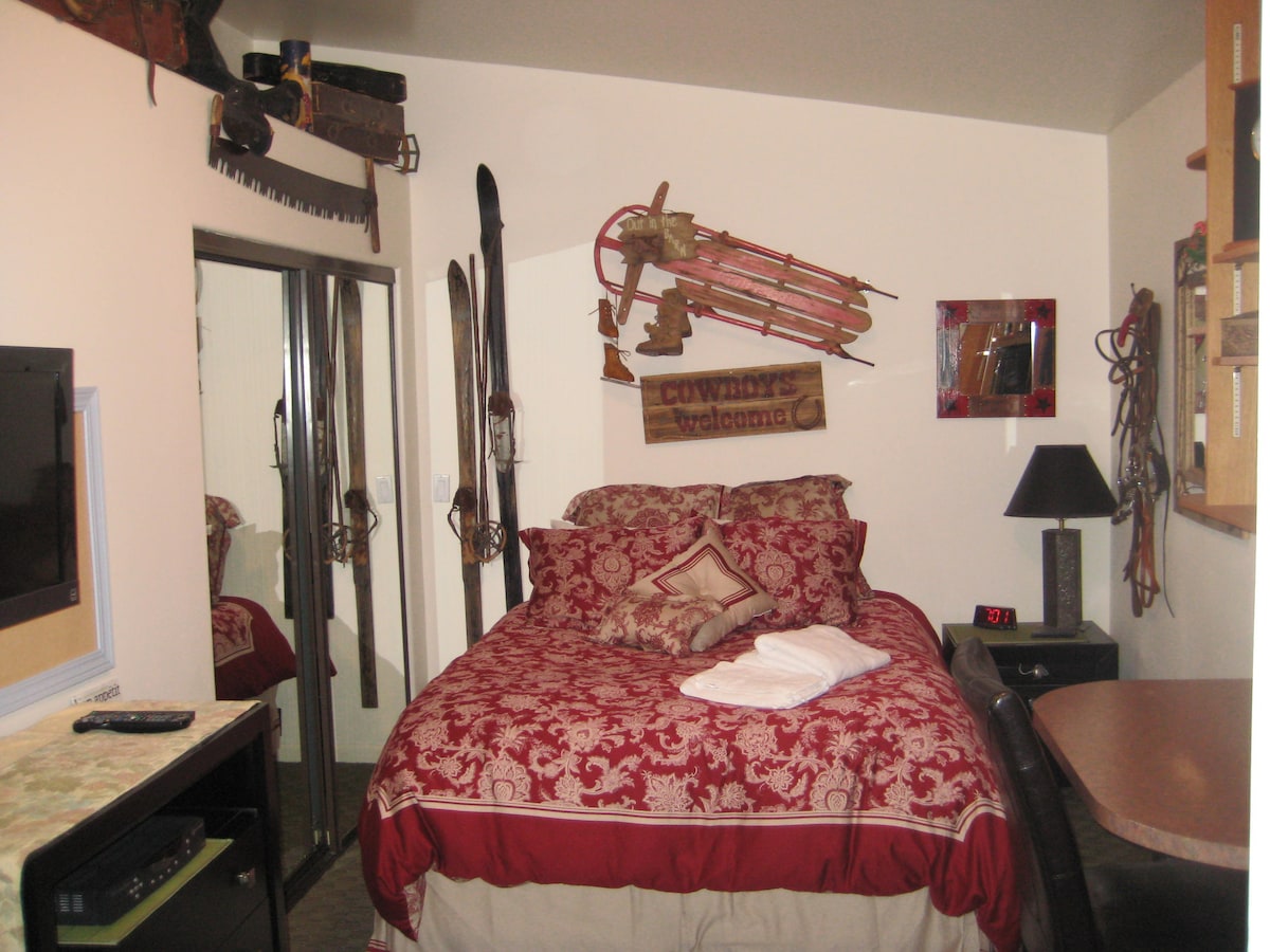 A spacious room is furnished with a queen bed dressed in a richly patterned red and cream coverlet. Western decor, including vintage skis and a sled, adorns the walls. A mirrored closet and a desk with a lamp contribute to the functional layout.